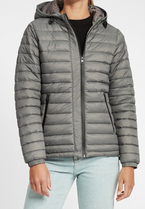 Personne portant une veste matelassée grise à capuche avec fermeture éclair frontale et poches zippées, associée à un jean bleu clair.