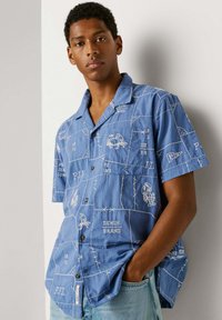 Junger Mann trägt ein blaues Kurzarmhemd mit weißen Stickmustern und Schriftzug, steht mit einer Hand in der Tasche seiner hellblauen Jeans.