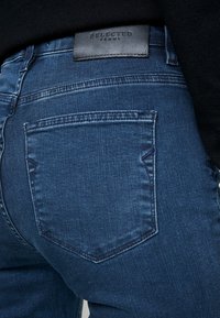 Jeans de mezclilla azul oscuro con una vista en primer plano del bolsillo trasero y la etiqueta de cuero. Presentan un sutíl bordado y una textura suave.