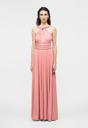 Sötét hajú nő, aki egy ujjatlan, rózsaszín, vállpántos maxi ruhát visel csomós elülső részlettel és ráncolt derékpánttal, egyszínű háttér előtt áll.