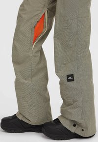 Grå mønstrede snebukser med lynlåseventiler, der afslører orange net. Har et logo patch og sorte sneboots ved kanten.