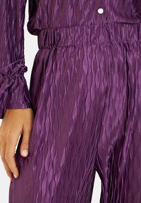 Fioletowa tkanina o fakturze, z falistym wzorem, elastycznym paskiem w talii i marszczonymi mankietami rękawów. Gładkie, błyszczące wykończenie.
