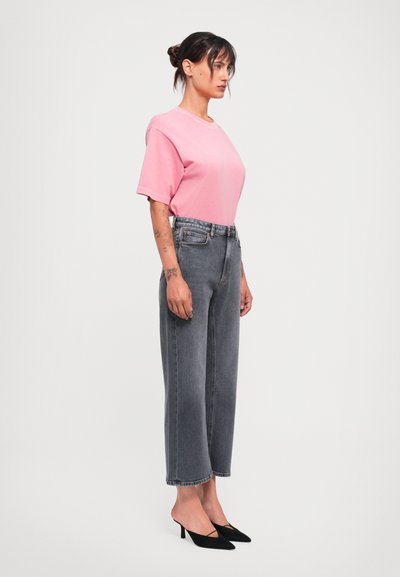 Woman with tattoos wears pink short-sleeve shirt, high-waisted faded gray jeans, and black kitten heel mules, standing sideways on white background.