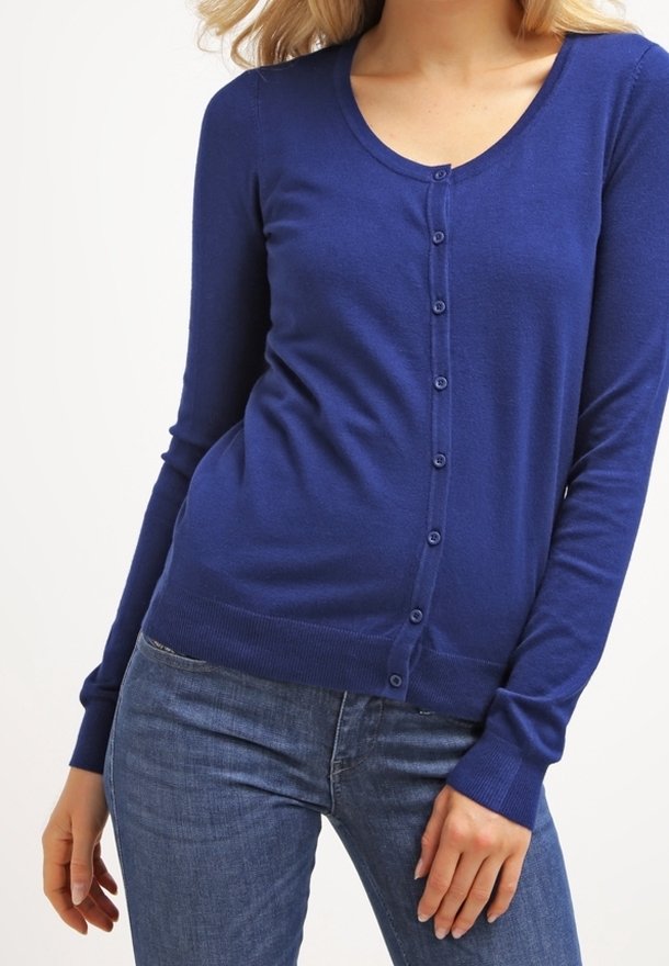 Femme portant un cardigan royal bleu ajusté à manches longues avec des boutons et un jean bleu, posant avec une main près de la cuisse.