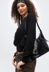 Νεαρή γυναίκα με σγουρά μαλλιά φοράει μαύρη μπλούζα με μακριά μανίκια, καφέ παντελόνι, μαύρη ζώνη, μαύρη κορδέλα και κρατάει μια μαύρη δερμάτινη τσάντα ώμου.