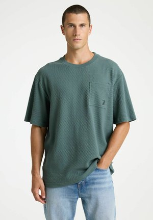 Jonge man met kort haar draagt een gestructureerd groen oversized T-shirt en lichtblauwe spijkerbroek, met één hand in de zak, op een effen witte achtergrond.