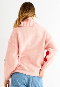 Pink knitted sweater with a ribbed turtleneck and red bow detail on the sleeve. Loose fit, textured fabric, and casual style.