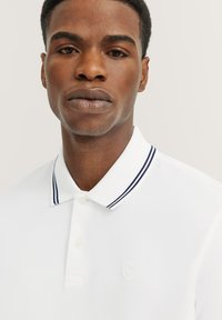 White polo shirt with a textured fabric, featuring a two-button placket and a collar accentuated by navy and white stripes. Subtle logo detail.