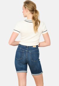 Denim šorti ar salocītām malām, tumšs tonis, piecu kabatu dizains un maiga ādas etiķete aizmugurējā viduklī. Īsām piedurknēm balta blūze ar melnu apdari.