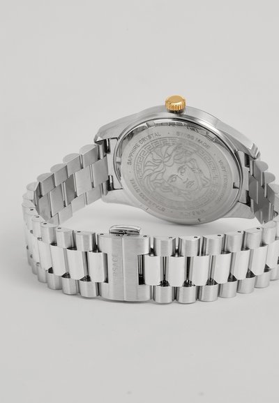 Montre-bracelet en métal argenté avec un bracelet à maillons, une couronne en or et une tête de Méduse gravée au dos.
