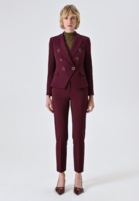 Abito bordeaux a doppio petto con dettagli in anelli argentati, vestibilità su misura, abbinato a pantaloni aderenti e tacchi in pelle lucida nera.