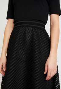 Schwarzer Rock mit einer strukturierten, wellenförmigen Musterung, ausgestattet mit einem breiten Bund und dekorativen Akzenten entlang des Saums. Glattes Material, figurbetonte Silhouette.