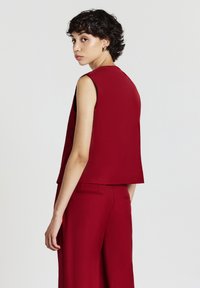 Donna con capelli ricci corti, in top senza maniche rosso e pantaloni rossi abbinati, che guarda oltre la spalla su uno sfondo semplice.