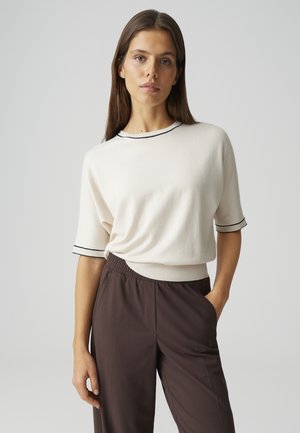 Donna con capelli castano lisci che indossa un maglione bianco a maniche corte con bordi neri e pantaloni a vita alta di colore marrone scuro, in piedi con una mano in tasca.