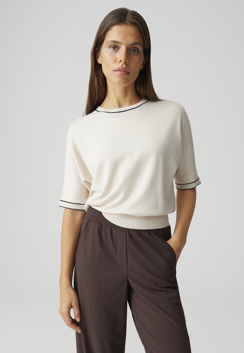 Femme aux cheveux bruns et raides portant un pull blanc à manches courtes avec bordure noire et un pantalon taille haute marron foncé, debout avec une main dans la poche.