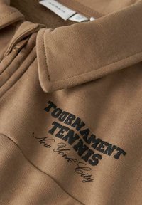 Smėlio spalvos polo marškinėliai su apykakle, ant kurių yra juodas siuvinėtas užrašas „TOURNAMENT TENNIS“ ir „New York City“. Matyti minkštos audinio tekstūros.