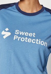 Zila īsām piedurknēm krekls, izgatavots no mīksta auduma, ar apaļu kakla izgriezumu. Priekšpusē redzami balti uzraksti "Sweet Protection". Tumši akcenti uz pleciem.