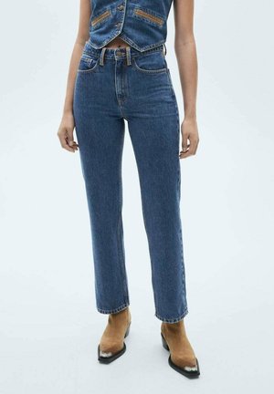 Kvinde iført højtaljede blå jeans med lige ben, en blå denimvest og brune ankelstøvler med spids tå og sølvspidser.