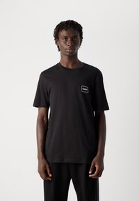 Ung mann med korte dreadlocks iført en svart HUGO t-skjorte og svarte bukser, står mot en ensfarget lys bakgrunn.