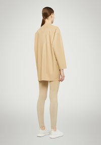 Oversized beige faux leather top with dropped shoulders, paired with metallic gold leggings and white sneakers, viewed from the back.