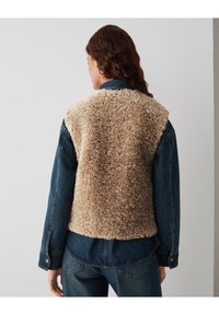 Personne portant un gilet en fausse fourrure bouclée beige sur une veste en denim bleu et un jean, de dos contre un fond uni.