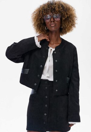 Femme aux cheveux bouclés portant des lunettes de soleil surdimensionnées, une veste noire à boutons et une jupe, ainsi qu'une blouse blanche avec un col noué.