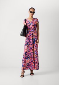 Robe maxi au motif paisley vibrant en rose et violet. Fabriquée en matériau doux avec des manches courtes, un décolleté en V et une taille cintrée. Accessoire de sac à main noir.