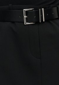 Μαύρη δερμάτινη ζώνη με ασημί ορθογώνια αγκράφα, φορεμένη πάνω από μαύρο υφασμάτινο παντελόνι και μαύρο ριμπ τοπ.