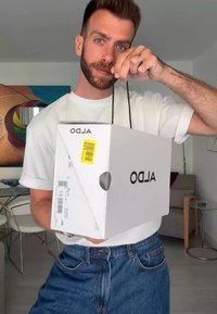 White rectangular box with a black logo, held by a person. Features a yellow price tag and black paper handles. Casual indoor setting.