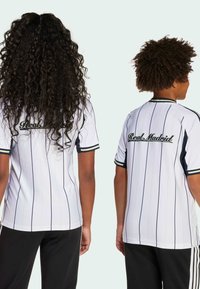Maglia bianca con strisce verticali nere, maniche corte e colletto tondo. "Real Madrid" è ricamato in nero sulla schiena.