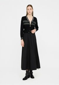 Cardigan zip nero con strisce orizzontali argento, abbinato a una gonna nera fluida e a stivali neri con tacco alla caviglia caratterizzati da dettagli in borchie.