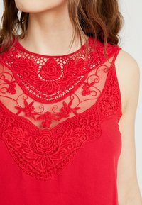Top senza maniche rosso con elaborata ricamatura in pizzo floreale rosso su una scollatura in rete trasparente, corpo in tessuto liscio e forma aderente.