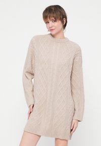 Rochie tricotată de culoare bej deschis, cu un model texturat în formă de diamant, tiv ondulat și mâneci largi pentru o potrivire lejeră.