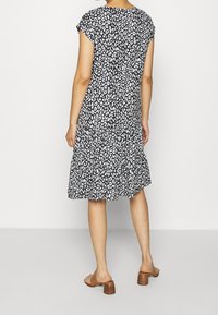 Kvinna klädd i en knälång klänning med svart och vitt leopardtryck samt bruna klacksandaler, stående med ryggen mot kameran.