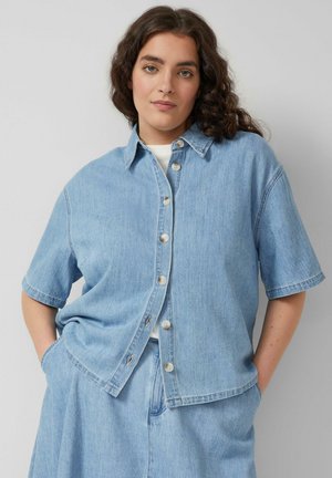 Kobieta o kręconych ciemnych włosach, ubrana w jasnoniebieską, krótkorękawową koszulę jeansową i dopasowaną spódnicę jeansową, ręce w kieszeniach, neutralne tło.