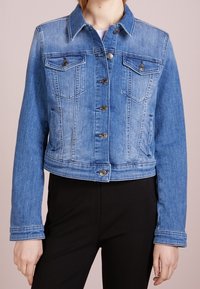 Modrá zkrácená džínová bunda s knoflíkovým zapínáním, dvěma náprsními kapsami, bočními kapsami, nošena přes černé kalhoty.