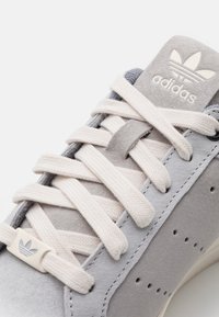 Grå mockaskor med vita snören, med en texturerad yta och en liten vit logotypdetalj nära snören.