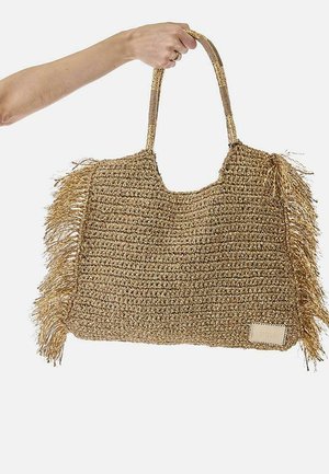 Mano sosteniendo un gran bolso tote de paja tejida con lados con flecos y dobles asas para el hombro sobre fondo blanco.