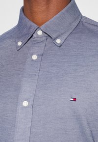 Camisa de botones azul claro hecha de tela tejida, con cuello con solapa, botones blancos y un pequeño logo con detalles en rojo, blanco y azul.
