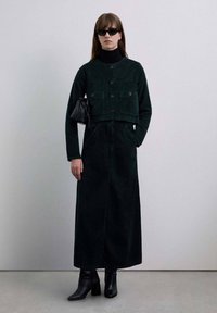 Chaqueta corta de pana verde oscuro con cuello alto y dos bolsillos en el pecho, combinada con una falda larga a juego hasta el suelo. Accesorios negros incluidos.