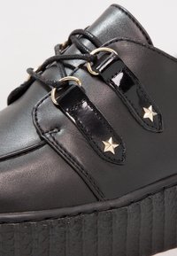 Chaussure à plateforme noire avec une semelle épaisse et texturée, des lacets noirs passés à travers des anneaux dorés et des clous décoratifs en forme d'étoile dorée sur des languettes noires brillantes.
