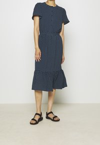 Žena v tmavě modrých midi šatech s krátkým rukávem a bílými puntíky, stojící na světlé jednobarevné podlaze a v černých sandálech na odkrytých prstech.