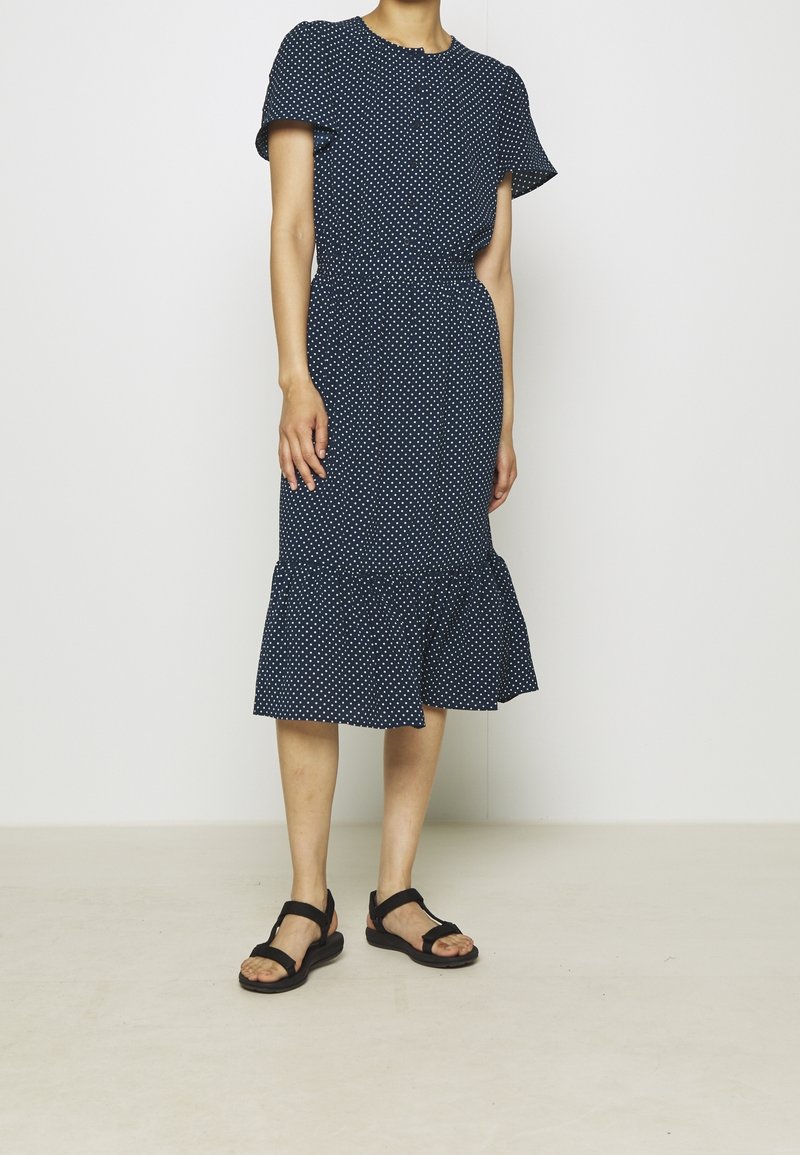 Žena v tmavě modrých midi šatech s krátkým rukávem a bílými puntíky, stojící na světlé jednobarevné podlaze a v černých sandálech na odkrytých prstech.