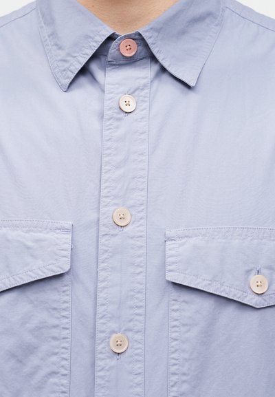 Φωτεινά γαλάζιο πουκάμισο με τσέπες μπροστά και κουμπιά σε αντίθεση, συμπεριλαμβανομένου ενός ξεχωριστού ροζ κουμπιού στο γιακά.