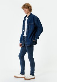 Chaqueta de mezclilla y jeans en azul oscuro, combinados con zapatillas blancas. La chaqueta cuenta con un bolsillo frontal y cierres de botón.