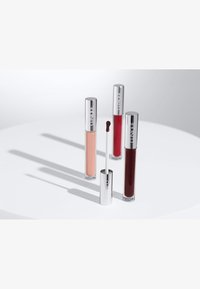 Four lip gloss tubes with reflective silver caps, featuring shades of nude, red, and deep burgundy, arranged on a white circular surface.