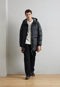 Daunenjacke aus schwarzem Stoff, mit Reißverschluss vorne, Kapuze und seitlichen Taschen. Kombiniert mit einem weißen Hemd und schwarzen Hosen, getragen mit dunklen Schuhen.