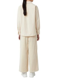 Camicia beige chiaro a maniche lunghe con bottoni e polsini arricciati, abbinata a pantaloni wide-leg dello stesso colore e sneaker bianche.