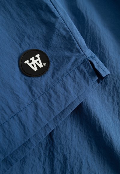 Blauer Stoff mit einer strukturierten Oberfläche, der einen schwarzen runden Aufnäher aufweist, der mit weißen Buchstaben "AW" geprägt ist, und einer sichtbaren Nahtdetail.