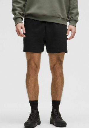 Ein Mann trägt einen grauen Pullover und schwarze Shorts. Er steht mit entspannten Händen an den Seiten, trägt schwarze Socken und Schuhe und steht vor einem neutralen Hintergrund.